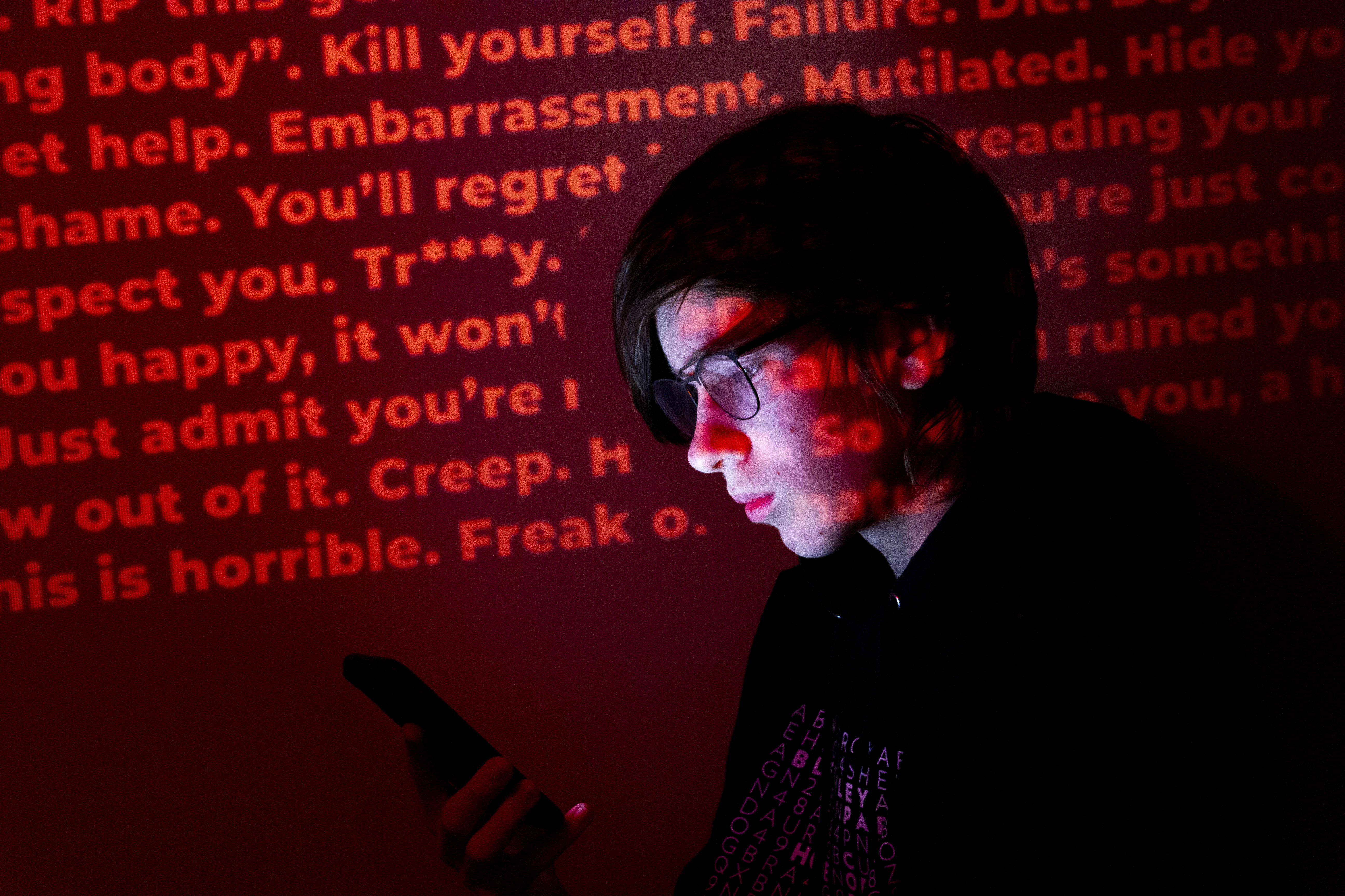 A person looking at a phone, with a melancholy expression. A wall projected with various insults and slurs targeted towards transgender people is behind them.