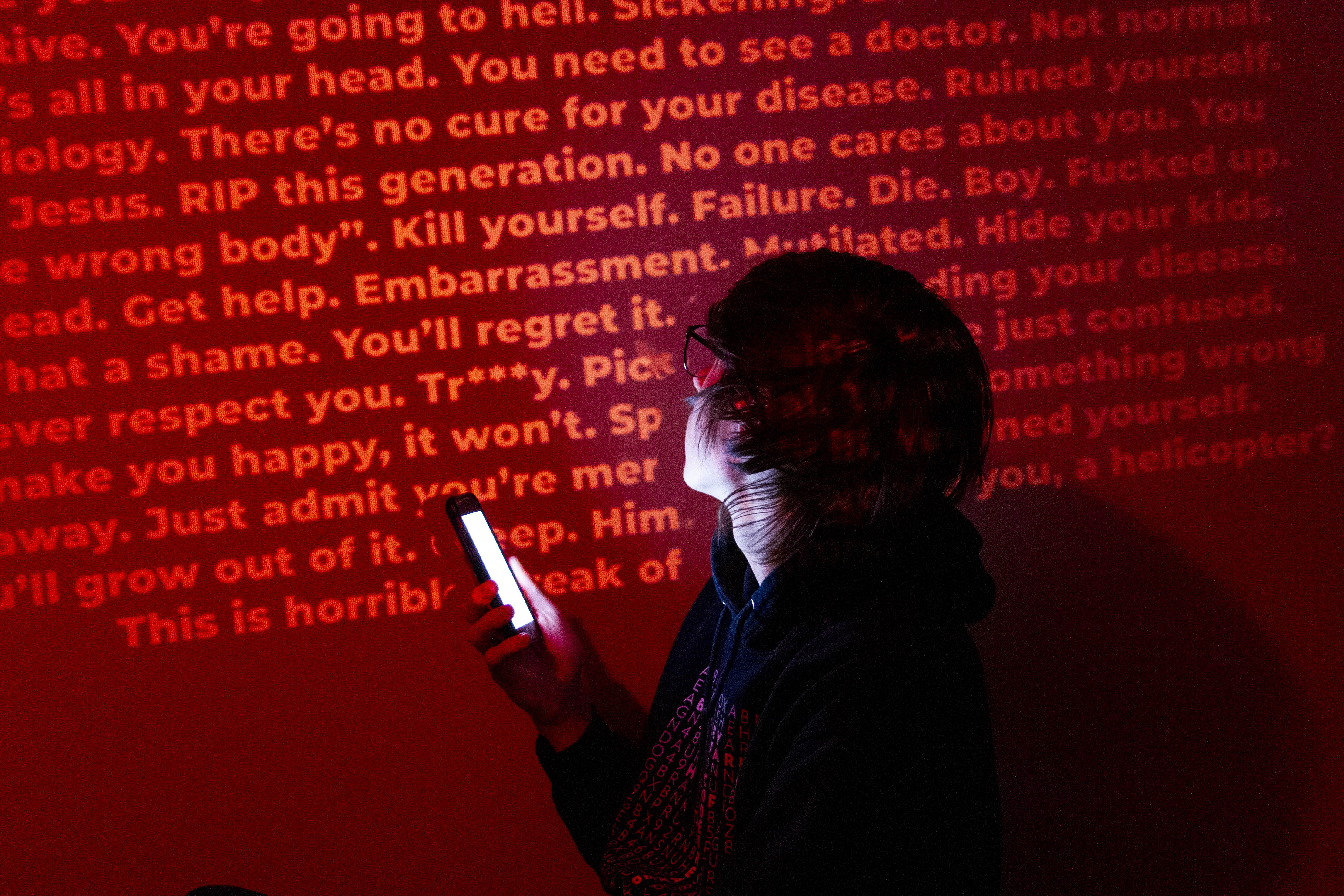  A person holding a phone, looking at a wall projected with various insults and slurs. 