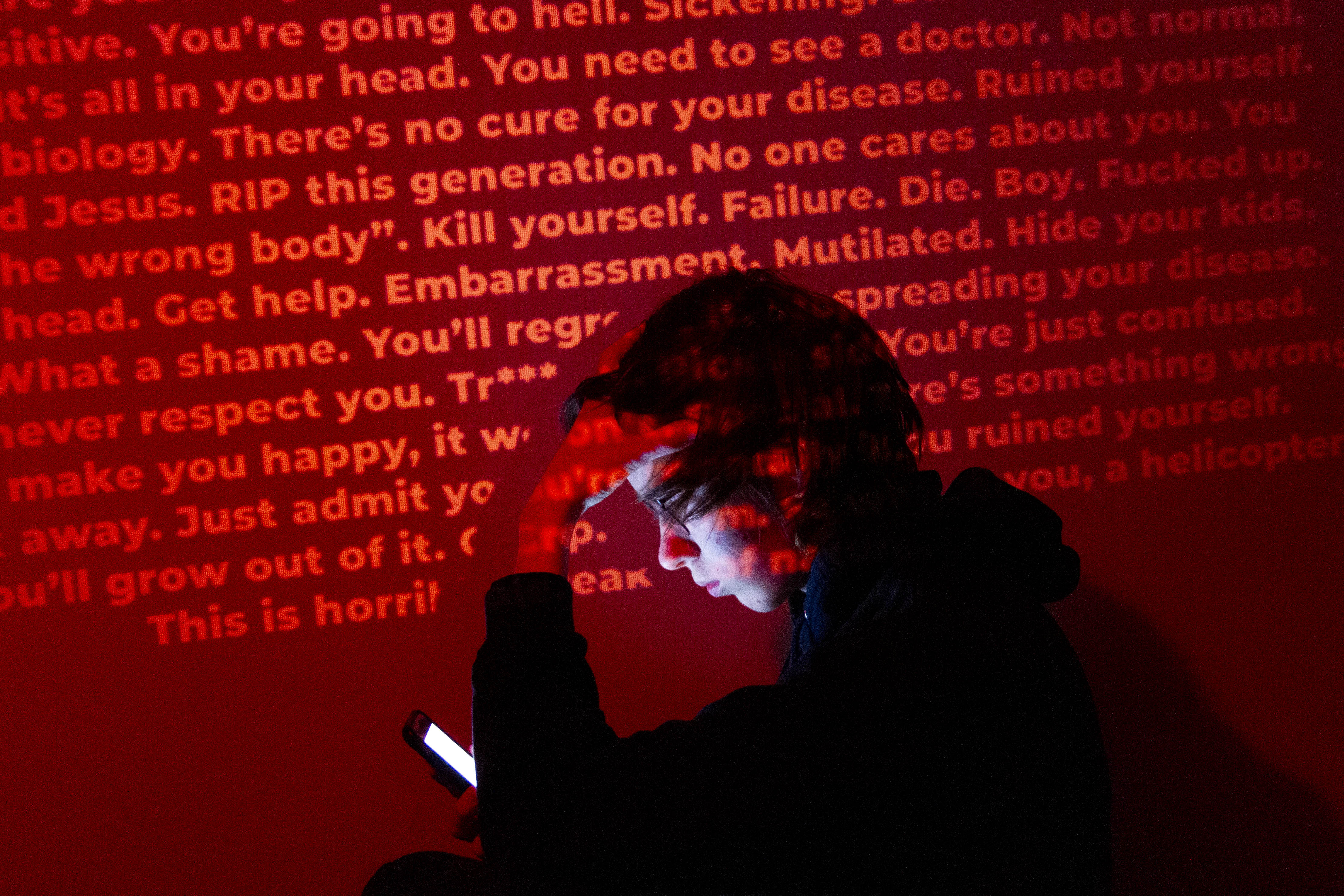 A person looking at a phone, with a melancholy expression. A wall projected with various insults and slurs targeted towards transgender people is behind them.