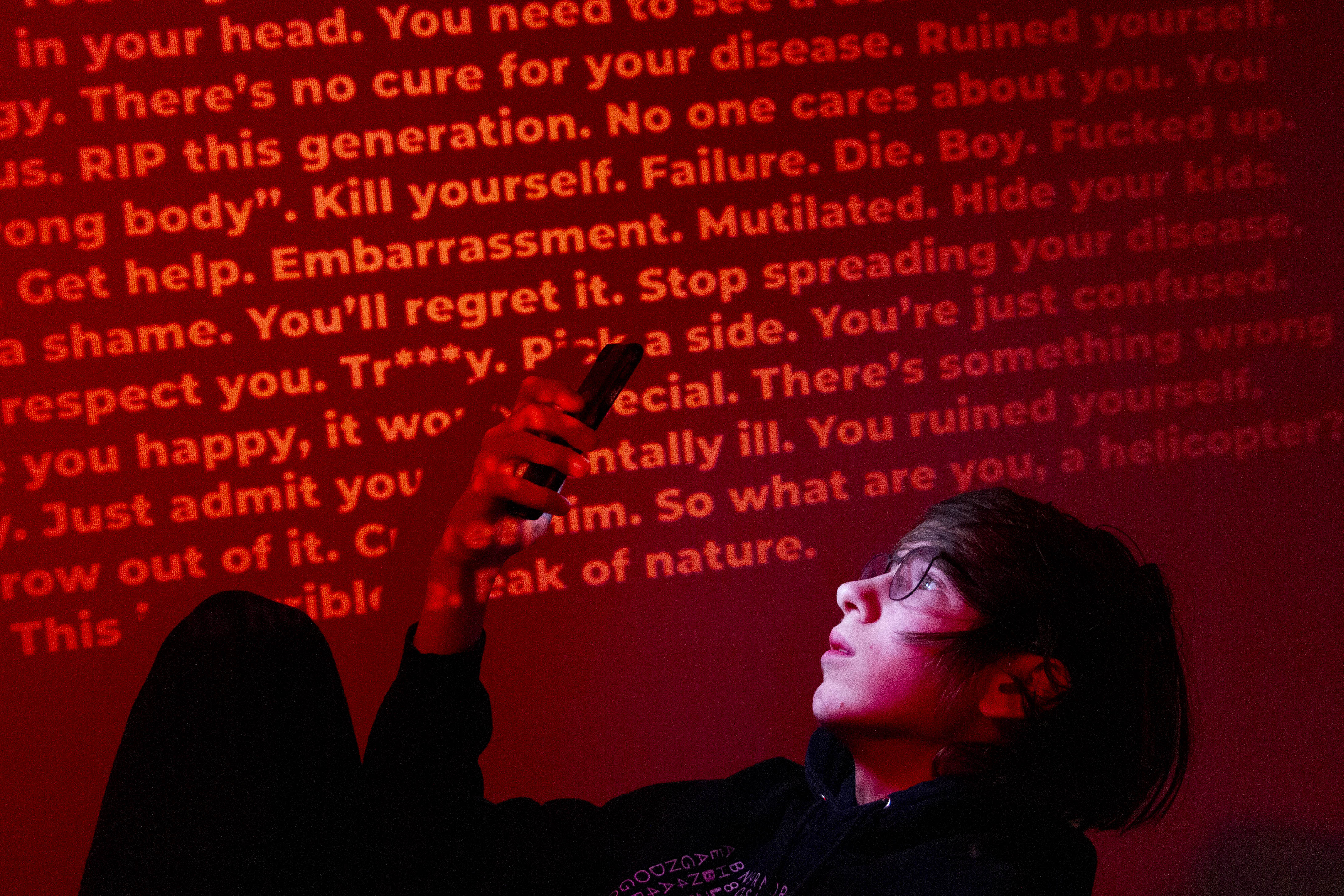A person looking at a phone. A wall projected with various insults and slurs is behind them.