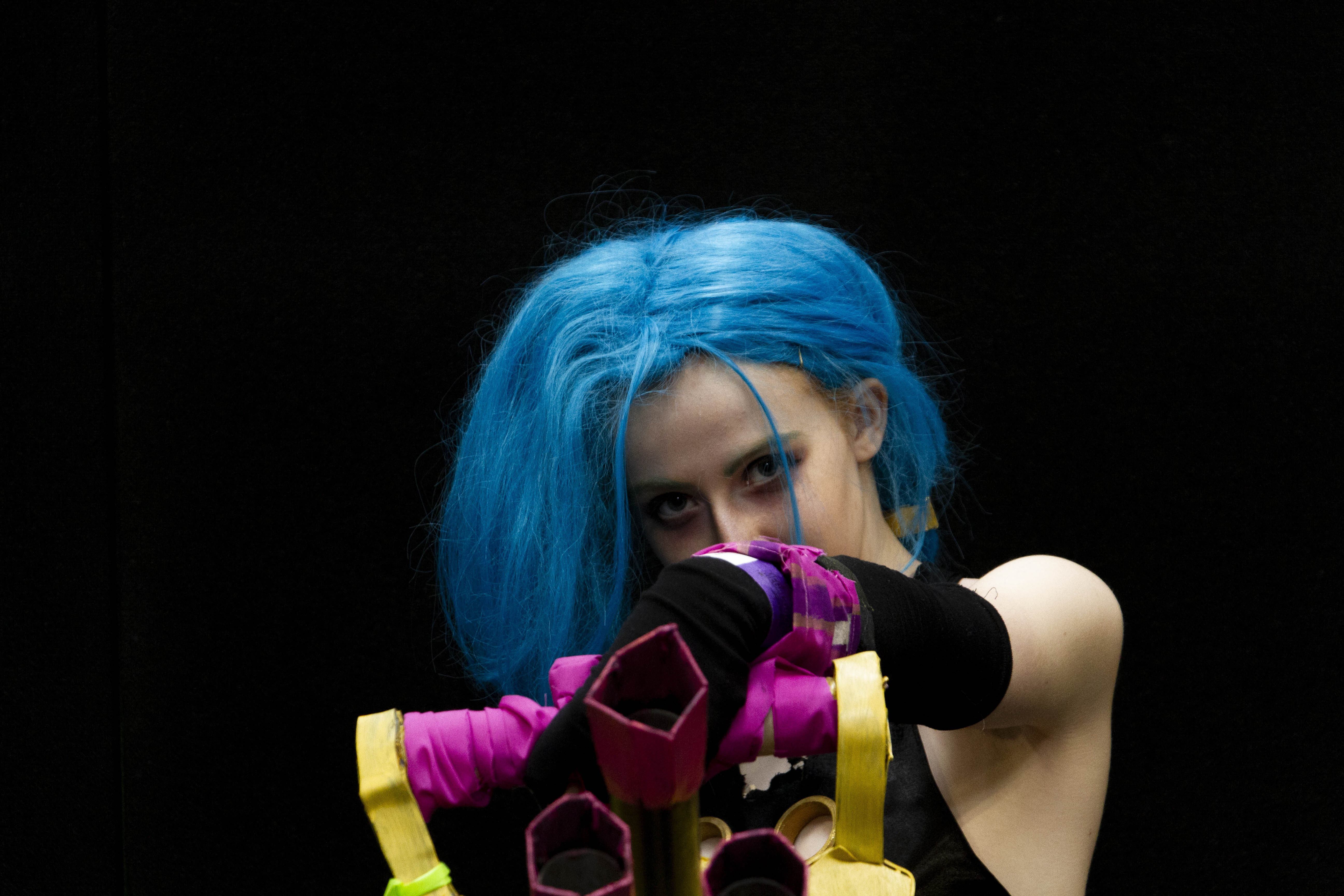 A person cosplaying Jinx from the TV Show Arcane. She is pointing a heavier, machine gun at the camera, looking at the camera with a dark expression.