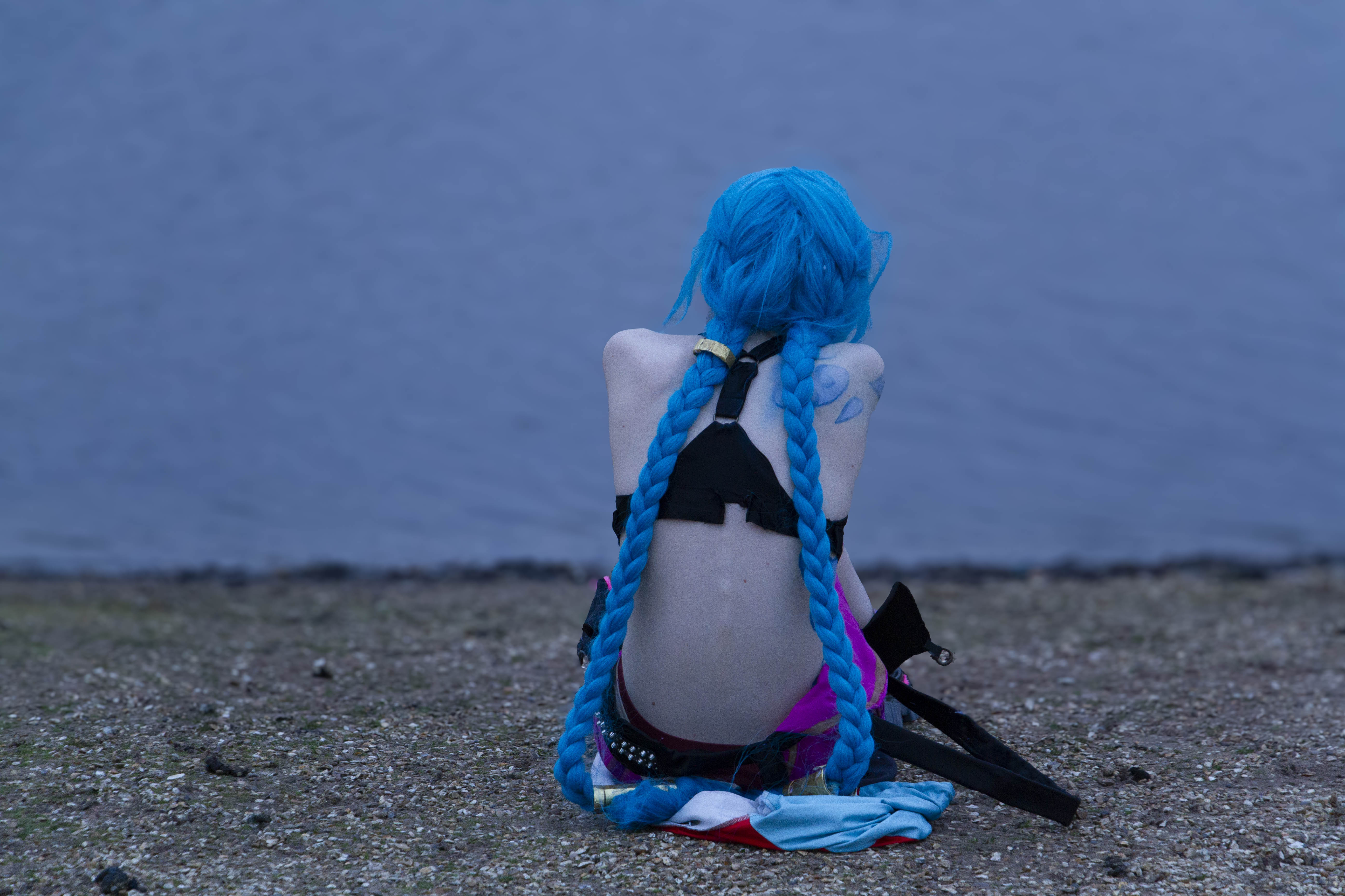 A person cosplaying Jinx from the TV Show Arcane. She is sitting on a rocky beach, with water in the background. She has her back turned towards the camera.