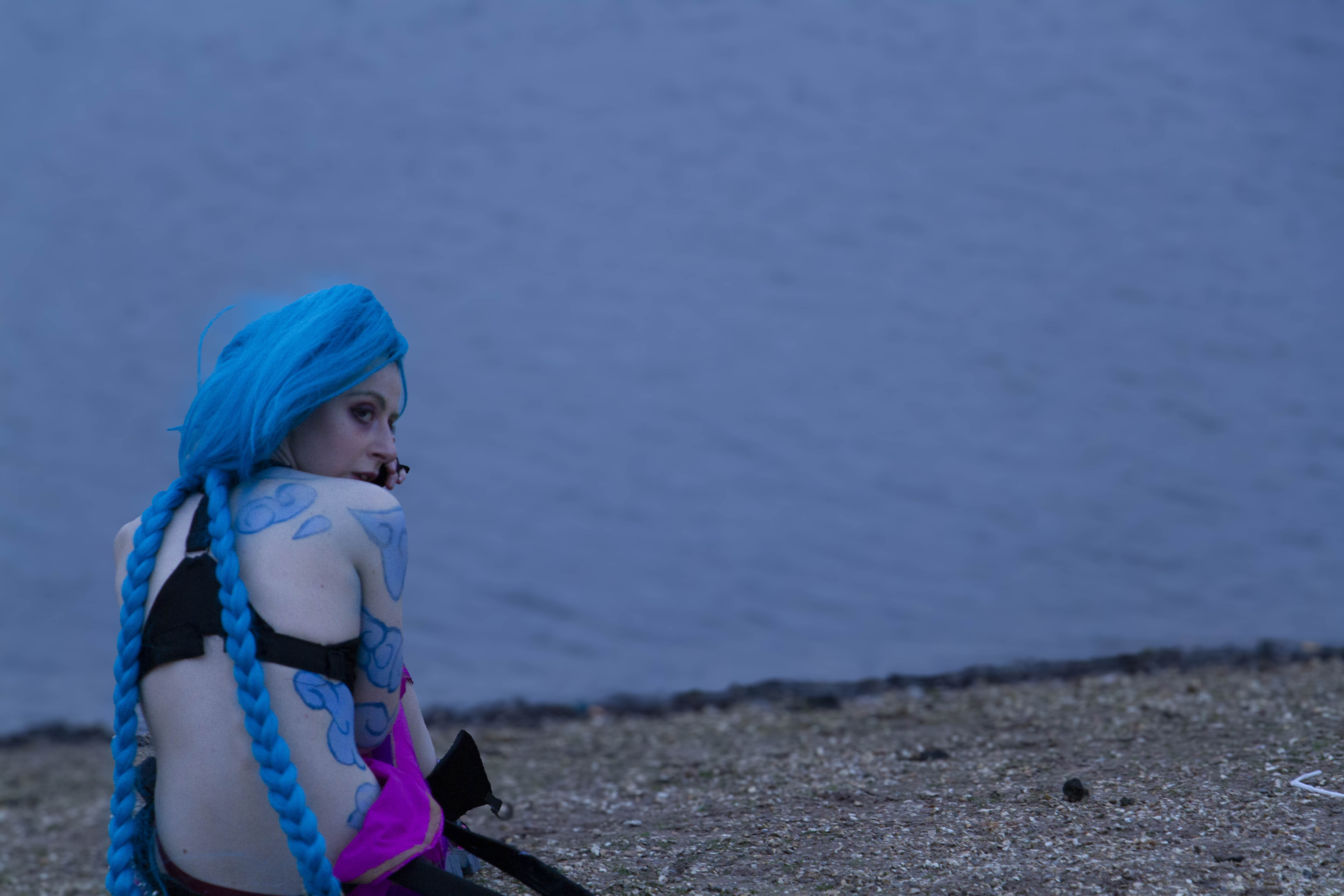  A person cosplaying Jinx from the TV Show Arcane. She is sitting on a rocky beach, with water in the background. She has her back turned towards the camera, looking back with a distressed expression 